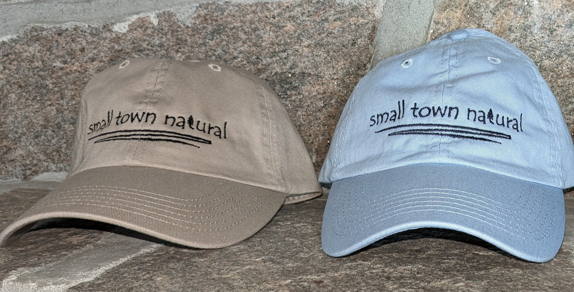 Hat 100% Cotton Small Town Natural  ('t' in natural is a tree embroidered graphic of same height as text) is embroidered in plain hand-printed text/font on front of your choice of sand or baby blue hathat in black thread (on black hat thread is light grey) with a thin zigzag line under text representing winding river / creek)