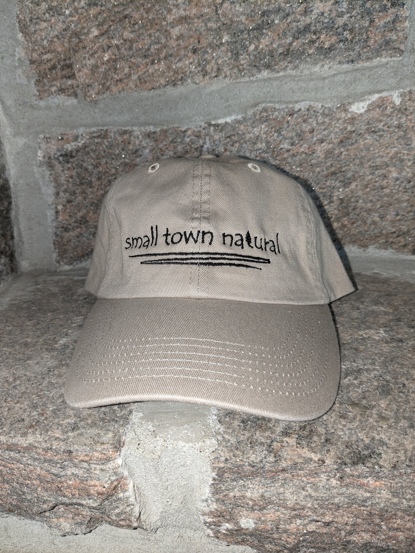 Hat 100% Cotton Small Town Natural ('t' in natural is a tree embroidered graphic of same height as text) is embroidered in plain hand-printed text/font on front of your choice of sand or baby blue hathat in black thread (on black hat thread is light grey) with a thin zigzag line under text representing winding river / creek)