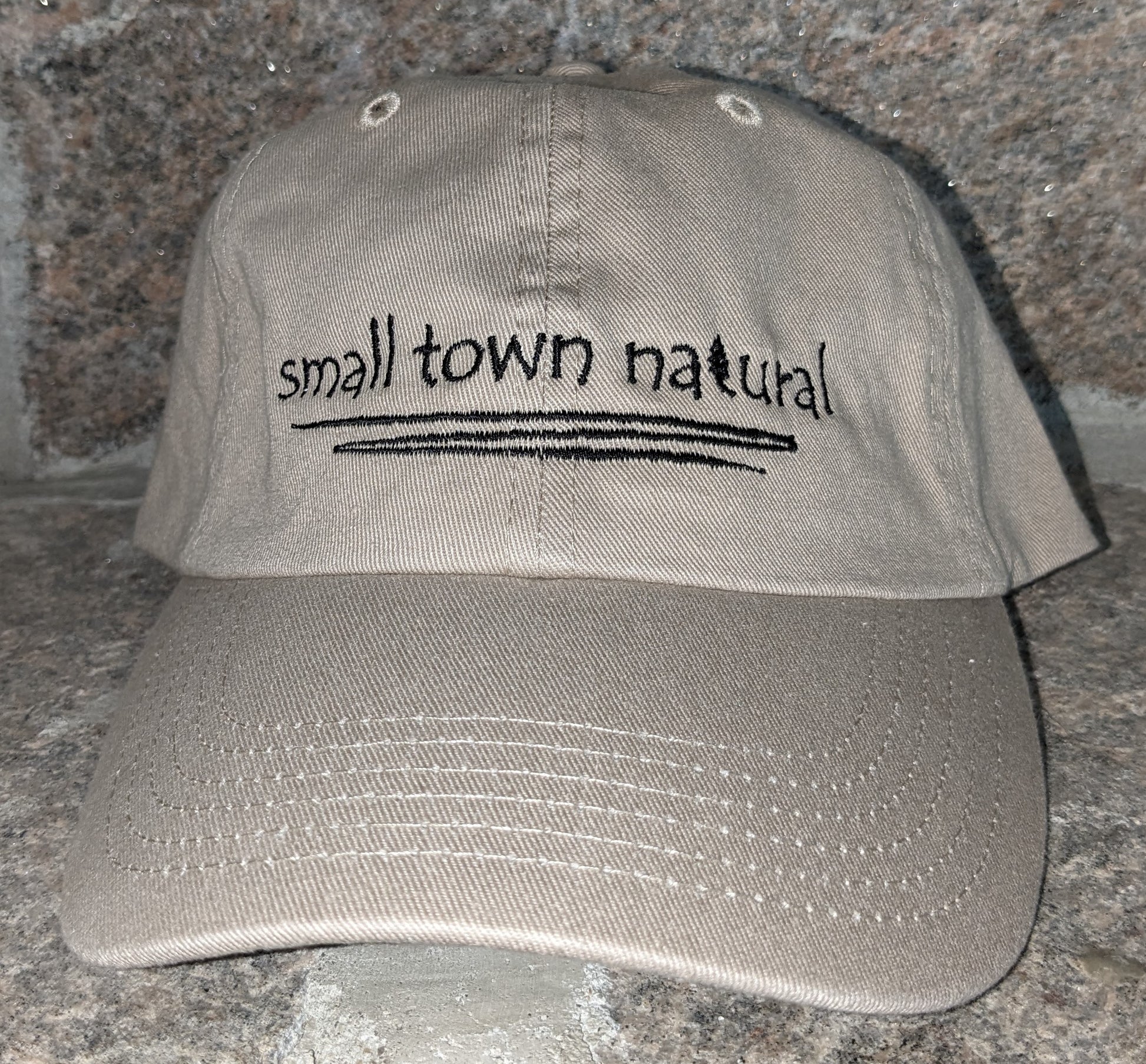 Hat 100% Cotton Small Town Natural ('t' in natural is a tree embroidered graphic of same height as text) is embroidered in plain hand-printed text/font on front of your choice of sand or baby blue hathat in black thread (on black hat thread is light grey) with a thin zigzag line under text representing winding river / creek)