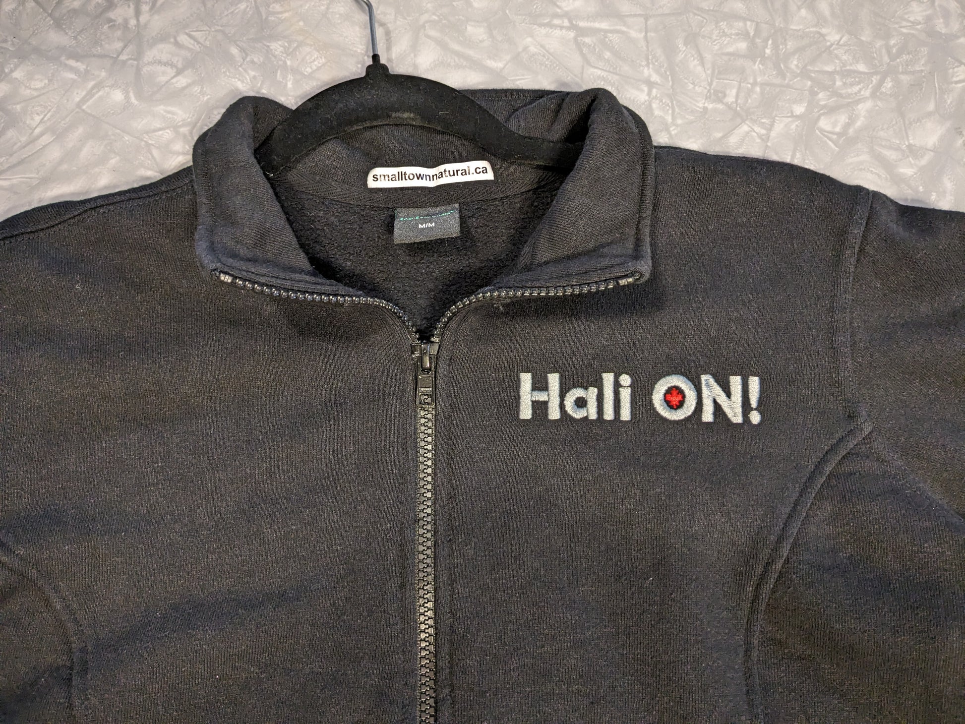 Small Town Natural original design of HALI ON! Hemp Fleece Track Jacket- Men's sizing, colour= black, (this is a close up of the HALI ON! embroidery in light grey with red maple leaf inside the 'O' )