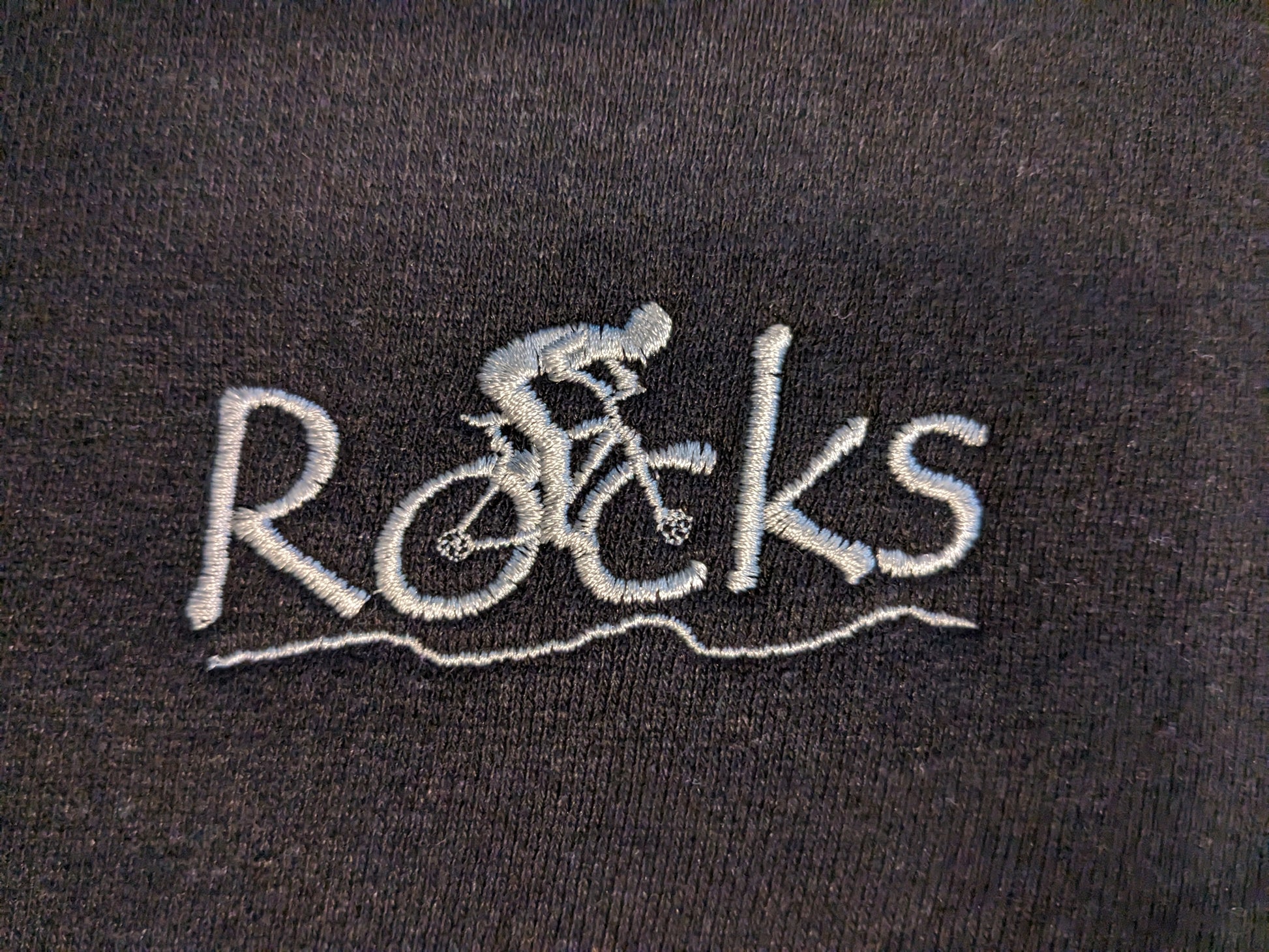 close up of embroidery on Rocks Hemp Fleece Track Jacket - Men's sizing. The embroidery spells Roads with 'oc' tires of bike with cyclist. Embroidery colour=silver light grey